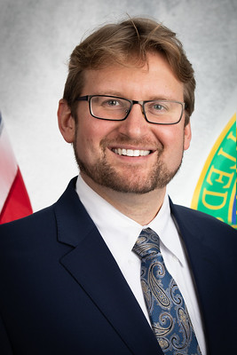A photo of Brian Anderson, a man with light brown hair, black glasses, and a navy blue suit jacket with a white button up shirt and a light blue print tie.