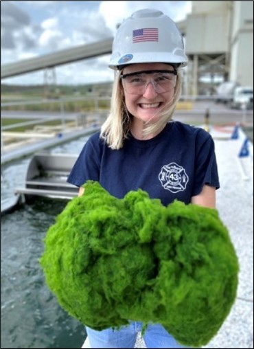 A blonde, Caucasian woman smiling and wearing a hard hat while holding algae.