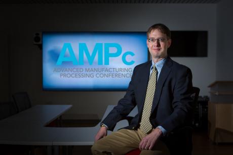 David Miller, a Caucasian man with short brown hair, wire frame glasses, a light blue button up shirt, navy suit jacket, yellow tie, and khaki pants sitting in front of a screen that reads "AMPc: Advanced Manufacturing Processing Conference"